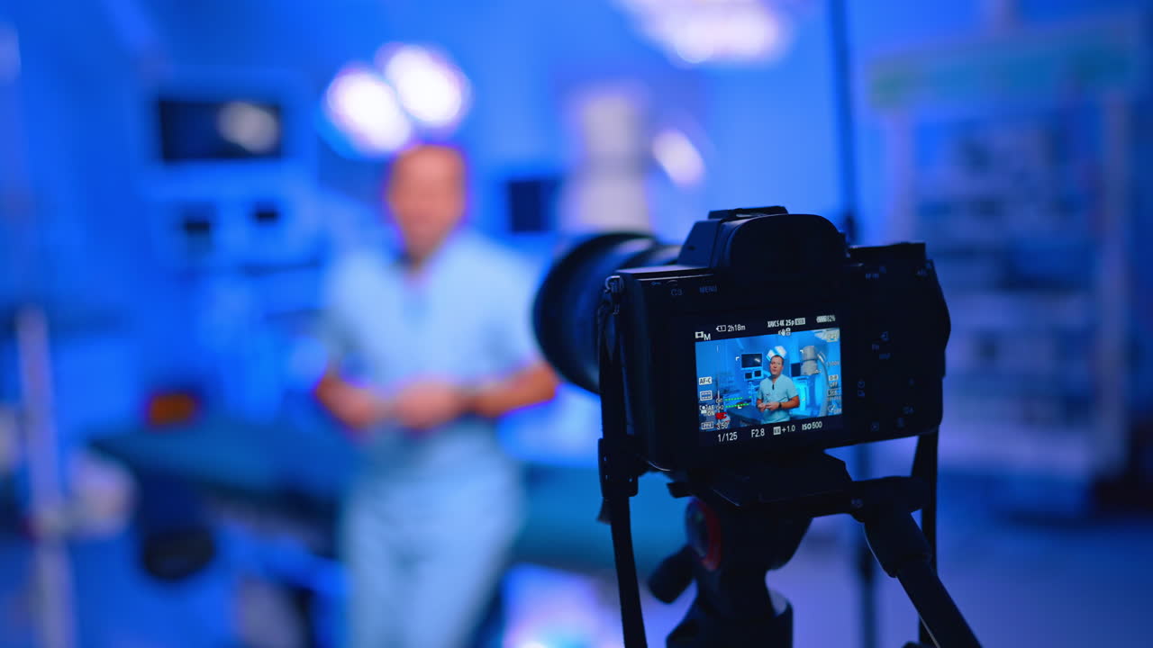 Telecamera ravvicinata riprende vlog di un medico in clinica blu. Lo schermo ravvicinato della telecamera mostra un medico in camice, sfondo sfocato di clinica blu, treppiede e luce LED.