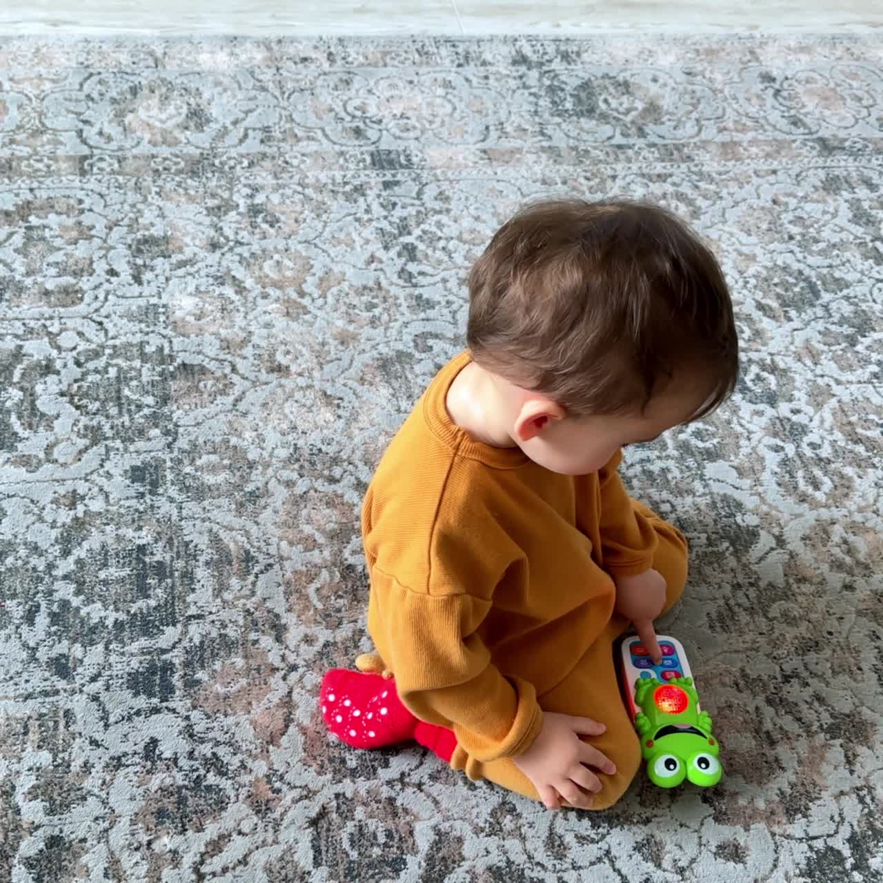 Baby boy in orange suit sitting on the floor playing with toy phone. Kid pressing the buttons on the toy. Top view
