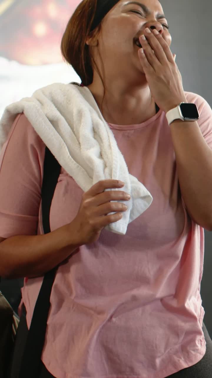 Vertical video: Yawning then laughing woman finishing workout at gym, with towel and light-pink top