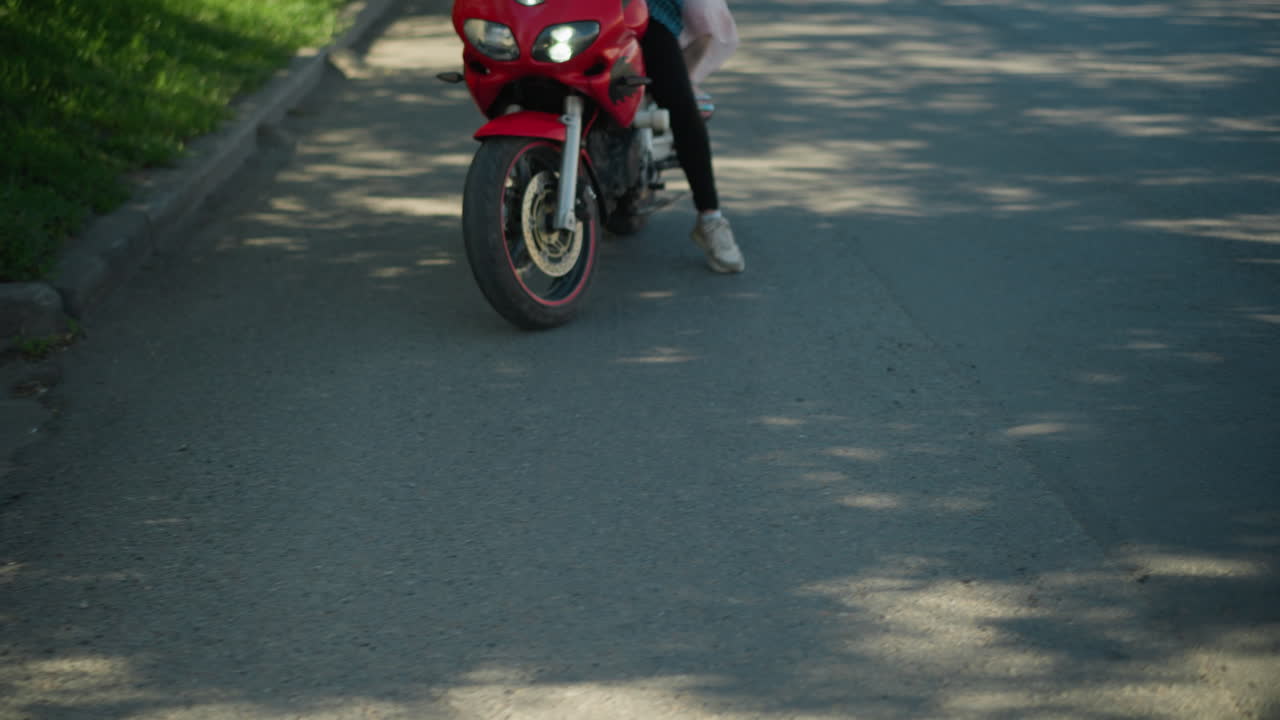две женщины на красном мотоцикле движутся по асфальтированной дороге, выложенной пышными зелеными деревьями, размытый объект виден рядом с камерой, в то время как тени и солнечный свет фильтруются через деревья