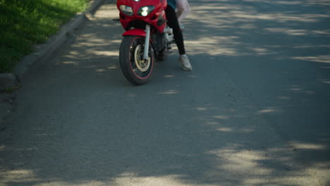 dos mujeres en una bicicleta eléctrica roja navegan por una carretera pavimentada bordeada de árboles verdes exuberantes, un objeto borroso es visible cerca de la cámara, mientras que las sombras y la luz del sol se filtran a través de los árboles