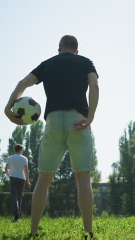 vista trasera de un hombre girando una pelota de fútbol a su alrededor mientras su hijo patea una pelota y un anciano la persigue