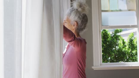 Kaukasische-ältere-Frau-Mit-Kaffeetasse,-Die-Zu-Hause-Die-Vorhänge-Des-Fensters-öffnet