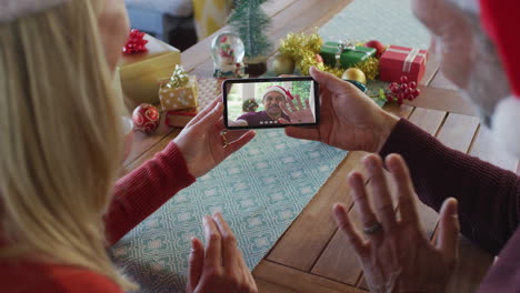 pareja caucásica con sombreros de santa usando un teléfono inteligente para una videollamada de navidad con un hombre en la pantalla