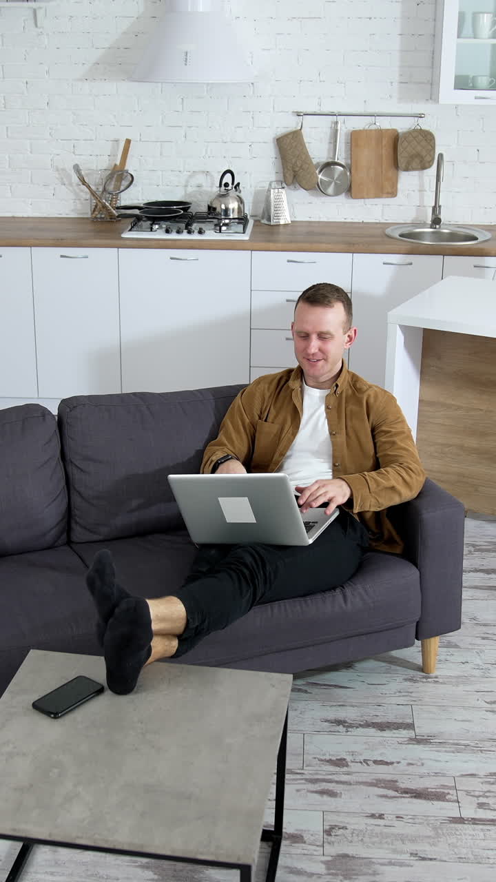 Young businessman working on a laptop. Modern kitchen background and man freelancer sitting in a couch with a laptop. Remote job. Vertical video