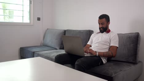 hombre barbudo sentado en un sofá escribiendo en una computadora portátil con una cara feliz