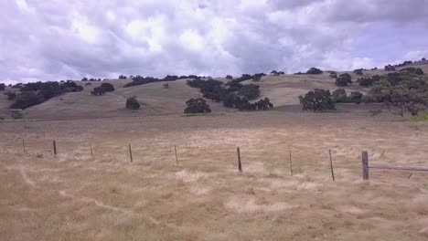 Slow-moving-drone-shot-flying-low-along-rustic-country-fence-to-reveal-foothills,-trees,-and-cloudy-blue-sky-in-background