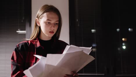 Young girl working at night in the office tired looking for paper looking through documents professionally looking at graphs standing at the window of a night city