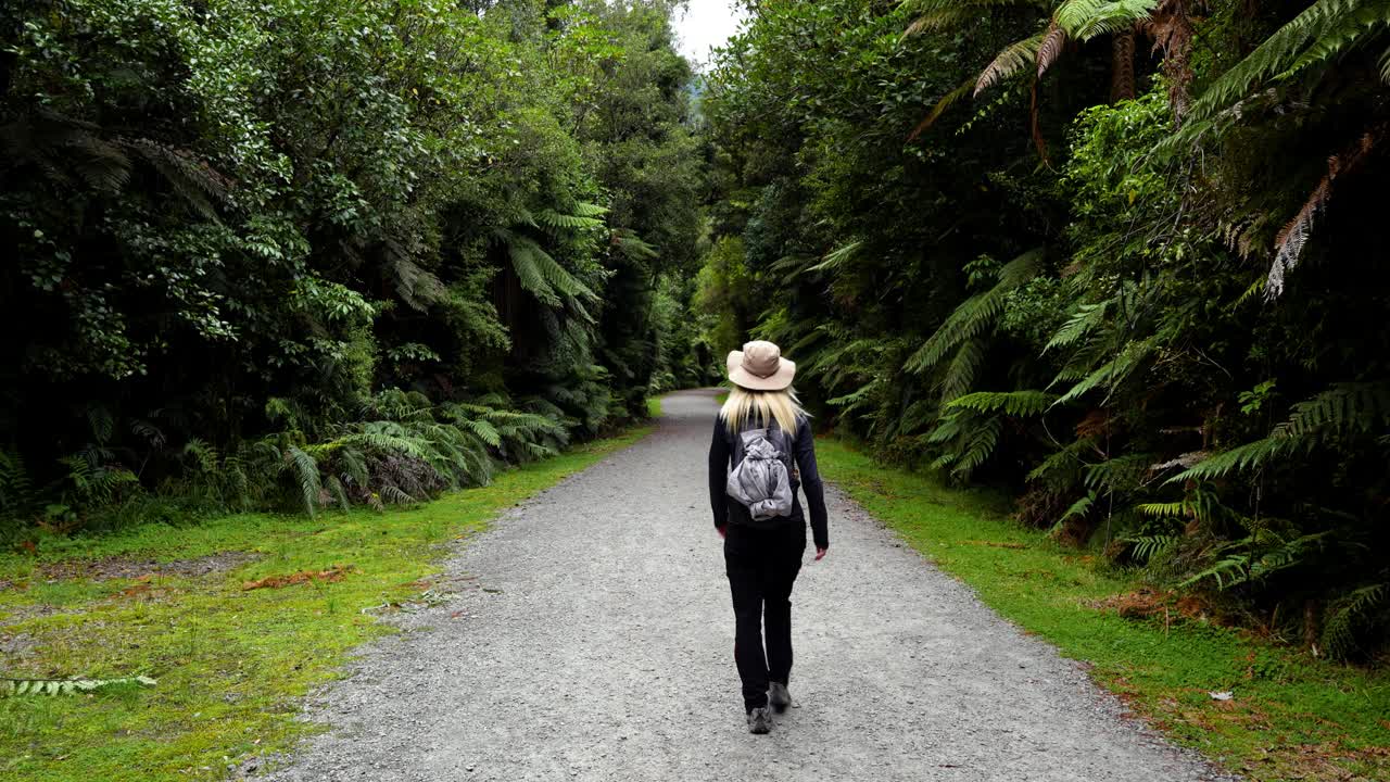 Blonde girl walking down the pathway through the lush Temperate Rainforest near Fox Glacier in New Zealand.
