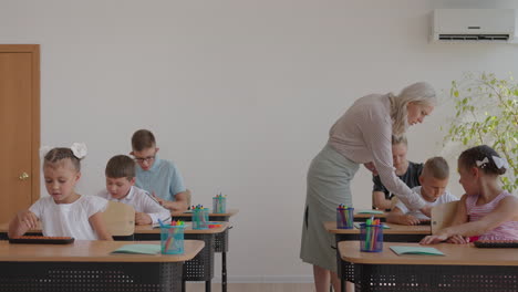 the-children-in-the-class-are-sitting-at-their-desks-and-the-teacher-walks-around-the-class-and-explains-the-topic-of-the-lesson.-Smart-Children-Learning-in-Friendly-Modern-Environment
