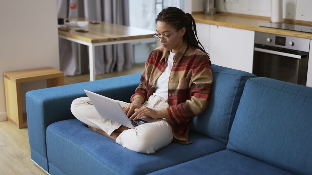 Woman with dreadlocks is working on new project and using laptop on couch in apartment room