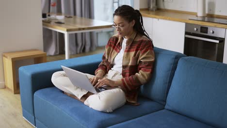 Woman-with-dreadlocks-is-working-on-new-project-and-using-laptop-on-couch-in-apartment-room