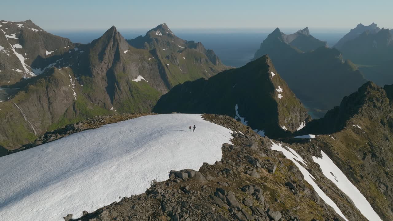 To personer står på den sneklædte, klippefyldte Munken-top i Lofoten, Norge, omgivet af tårnhøje granittoppe med vidtstrakt havudsigt under en klar blå himmel