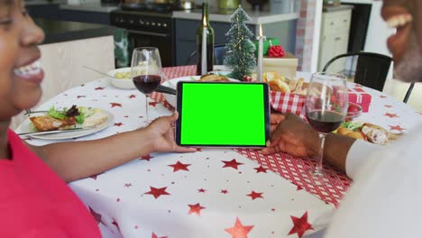 una pareja afroamericana sonriente con sombreros de santa haciendo una llamada de video de navidad de tableta, con pantalla verde