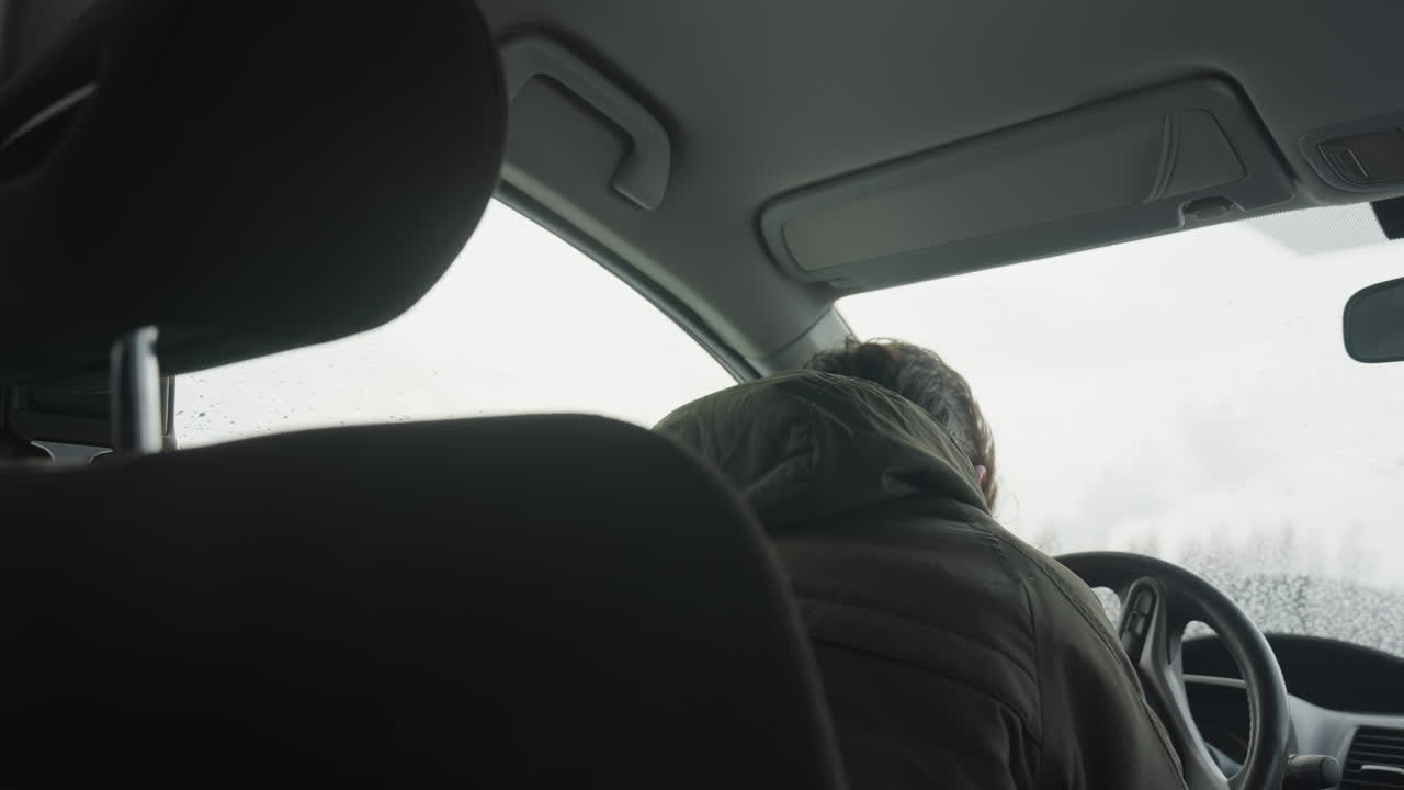close up of young boy in winter jacket inside vehicle closes door and adjusts seat while holding steering wheel with raindrops on window and blurred snowy landscape outside