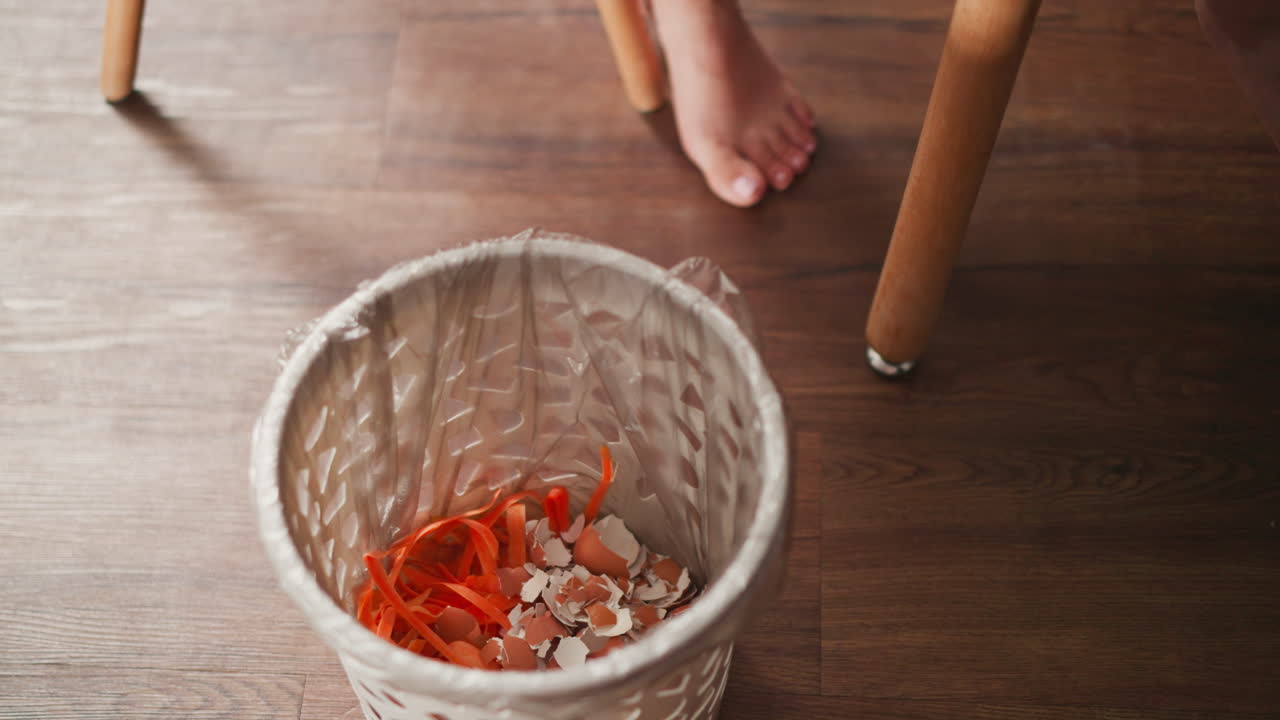 waste bin filled with bright carrot peels and broken egg shells on wooden floor with hand disposing leftover snacks from plate into bin beside wooden chair and visible bare foot