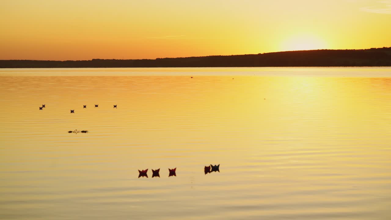 Beautiful picture of floating paper boats on the calm river at an orange sunset. Homemade origami ships on the evening water surface.