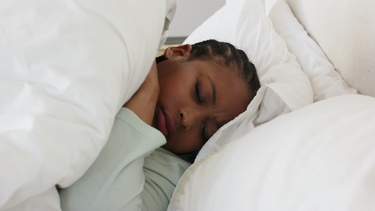 En afroamerikansk kvinde føler smerte i sengen, griber fat i dynen, masserer nakken og putter sig under dynen.