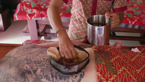 spa therapist uses a cloth to remove oil from the wax-lined container on the back of a patient enjoying a kati vasti treatment