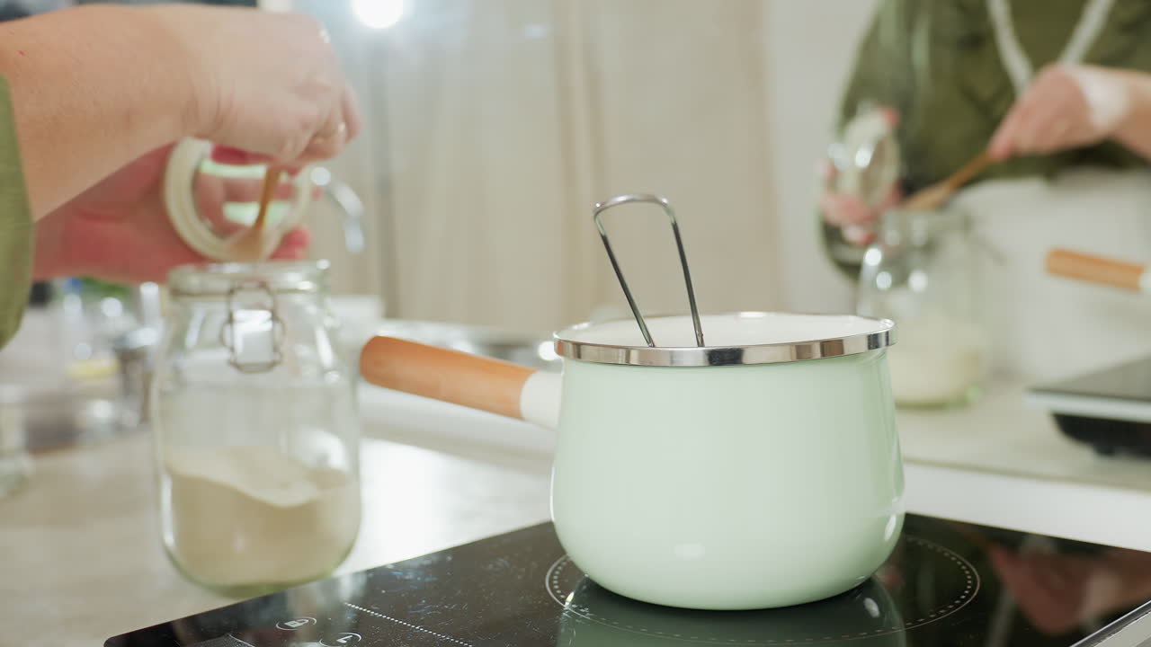 Chef adds salt into boiling pot using small wooden spoon and returns spoon to glass jar in bright kitchen with green cookware on electric stove during food preparation process