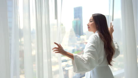 Eine-Hübsche-Frau-Im-Bademantel-Zieht-Die-Durchsichtigen-Vorhänge-In-Ihrem-Hotelzimmer-Zurück,-Um-Den-Blick-Auf-Die-Skyline-Einer-Modernen-Stadt-Freizugeben