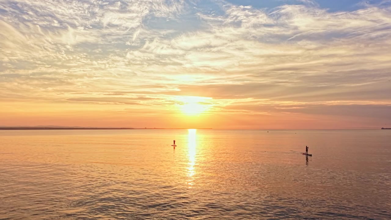 Paddleboarders enjoy sunset on calm water at the beach with a colorful sky in the background