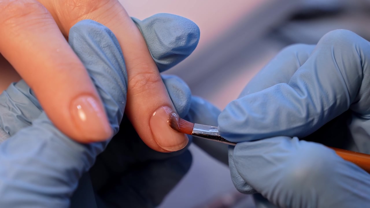 Foto em close-up em salão de uma designer de unhas aplicando gel construtor em uma unha anelar, destacando o nivelamento preciso do produto, o acabamento brilhante e o apoio profissional e cuidadoso da mão.
