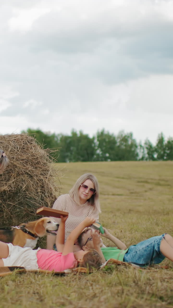 할머니가 딸에게 책을 건네주고, 어린 소녀는 동생에게 책을 건너주고, 이모는 가까이 앉아 가족이 넓은 농장에서 함께 시간을 즐기고, 개는 에 서 있습니다.
