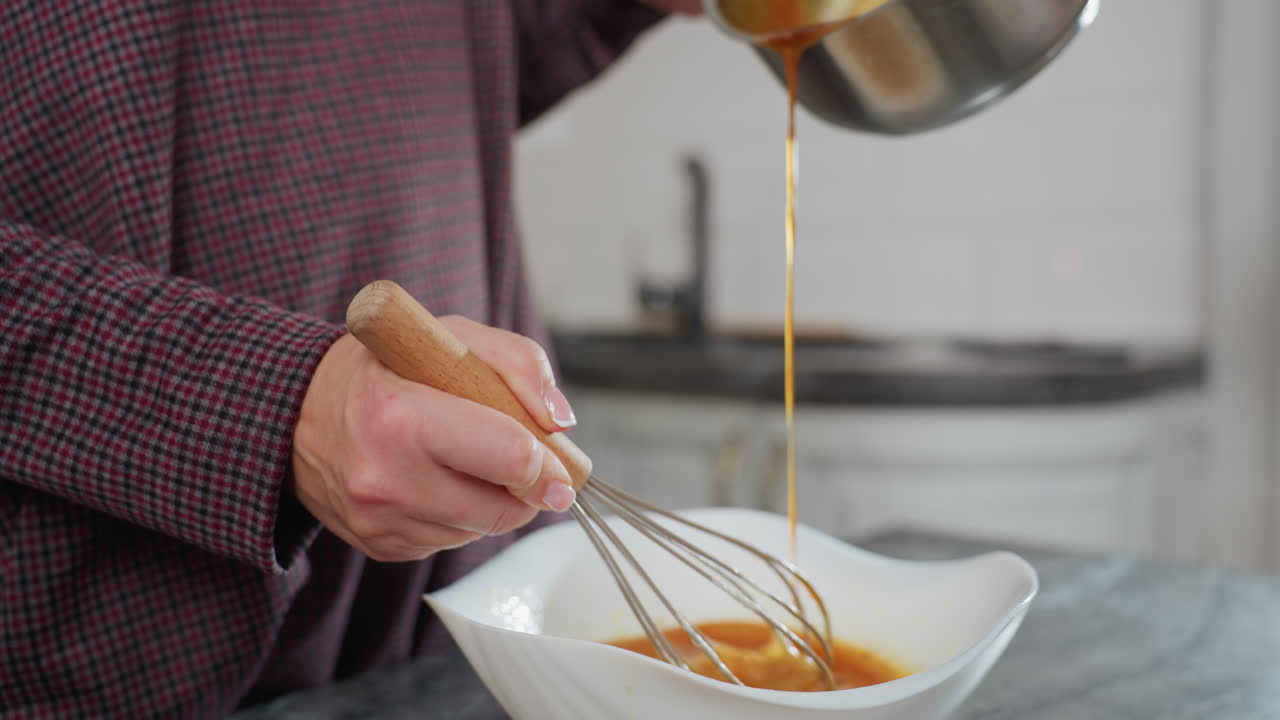 Close-up of cook pouring hot liquid into white bowl while stirring with whisk, hands actively mixing and adding ingredients in modern kitchen, warm soup blends with egg mixture for homemade dish