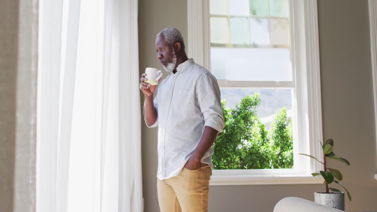 Anziano uomo afroamericano che beve caffè mentre guarda fuori dalla finestra a casa.