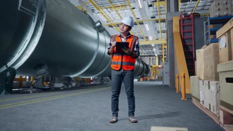 cuerpo lleno de ingeniero masculino asiático con casco de seguridad de pie en la fábrica de fabricación de tubos. tomando nota en la tableta y mirando a su alrededor