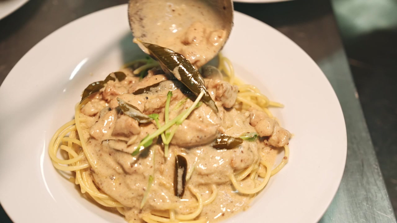 Ripresa macro in movimento di uno chef che usa un mestolo per versare una salsa ricca, densa e cremosa con pollo e foglie di curry su mucchi di spaghetti cotti su piatti bianchi.