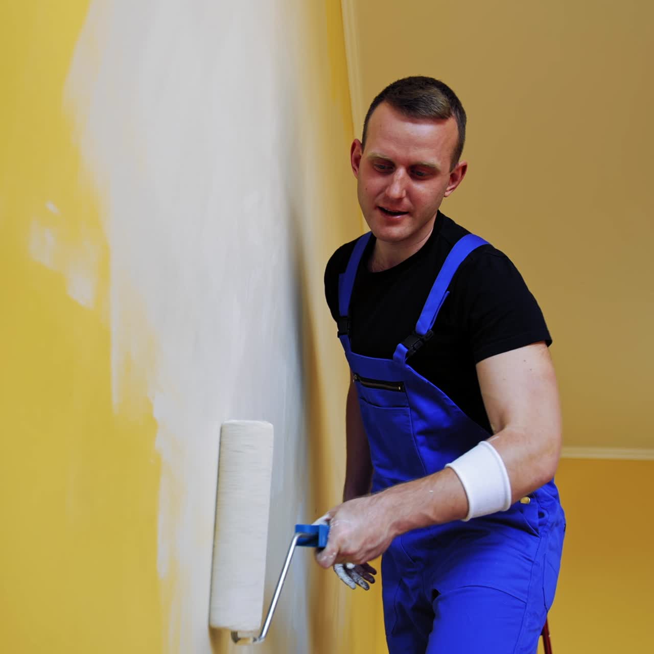Portrait of a painter in action. Young man builder in blue overalls decorating room with a paint roller in the apartment. Room makeover