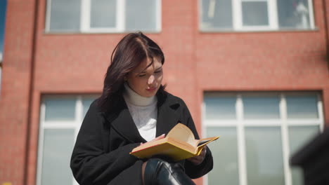 woman in black coat sitting outside, thoughtfully reading her book as sunlight illuminates her face, her hair moves gently in the breeze, with a brick building in the background