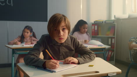 Schoolboy-writing-in-notebook-at-lesson.-Thoughtful-boy-holding-pen-at-mouth