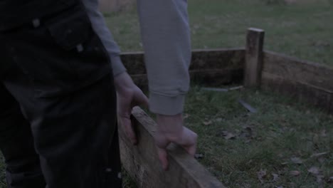 Young-man-wearing-hoodie-placing-plank-building-raised-garden-bed-at-dusk