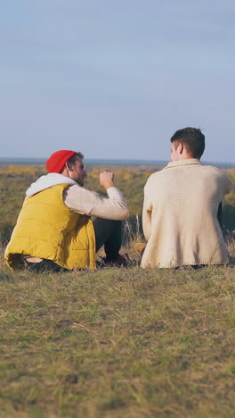 young men spend time sitting on hilly river bank at campsite in warm autumn evening backside view