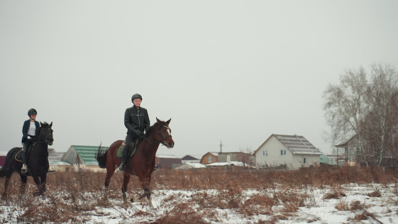 Coppia di cavalieri attraversa un freddo paesaggio invernale. Coppia caucasica a cavallo attraverso una campagna aperta e gelida. Due individui a cavallo che esplorano un vasto terreno invernale con lo skyline del villaggio.