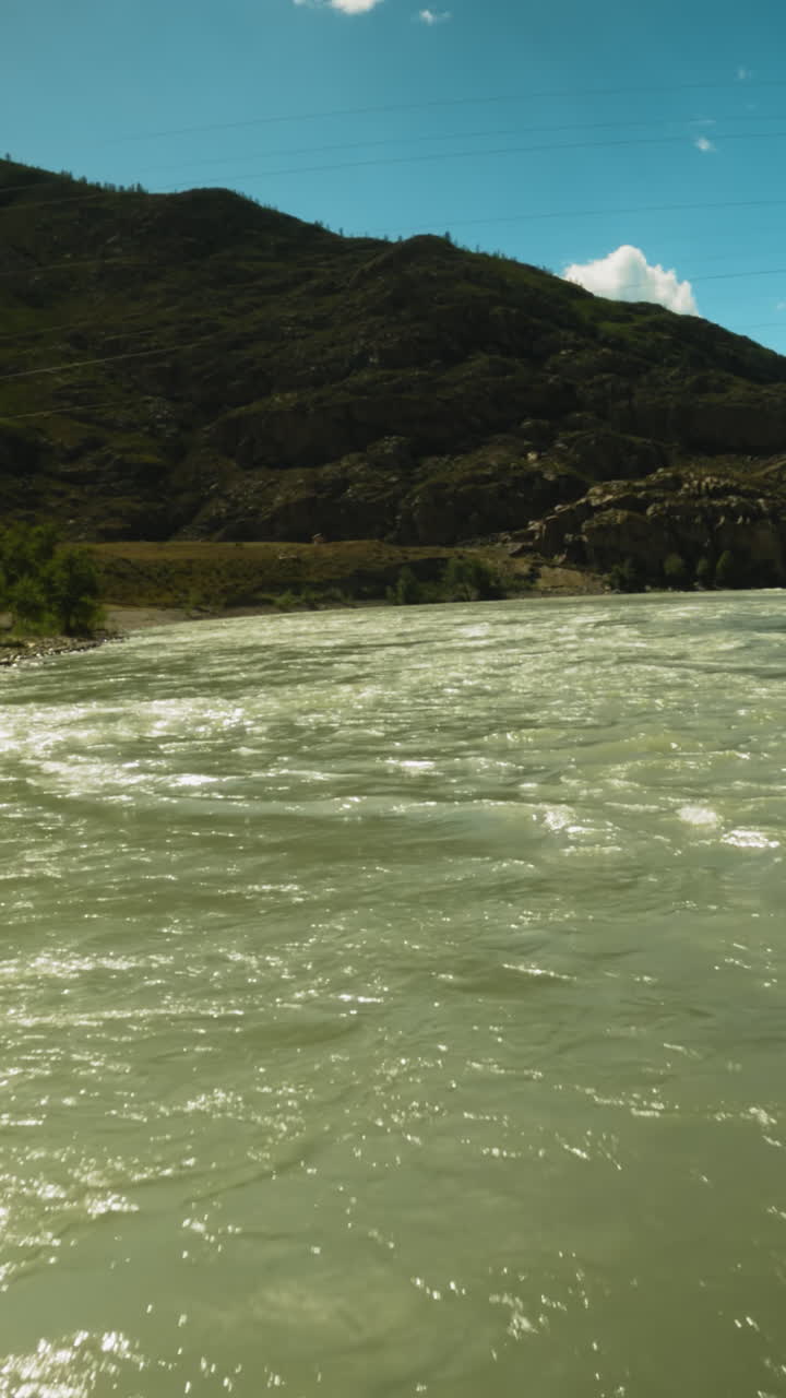 Mountain river rushes along wide course fpv. Fast untamed water stream glisten in bight sunlight against large mountain. Fresh creek at highland