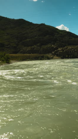 el río de montaña se precipita a lo largo de un amplio curso fpv. rápido arroyo de agua indomable brilla en la luz del sol de la bahía contra la gran montaña.