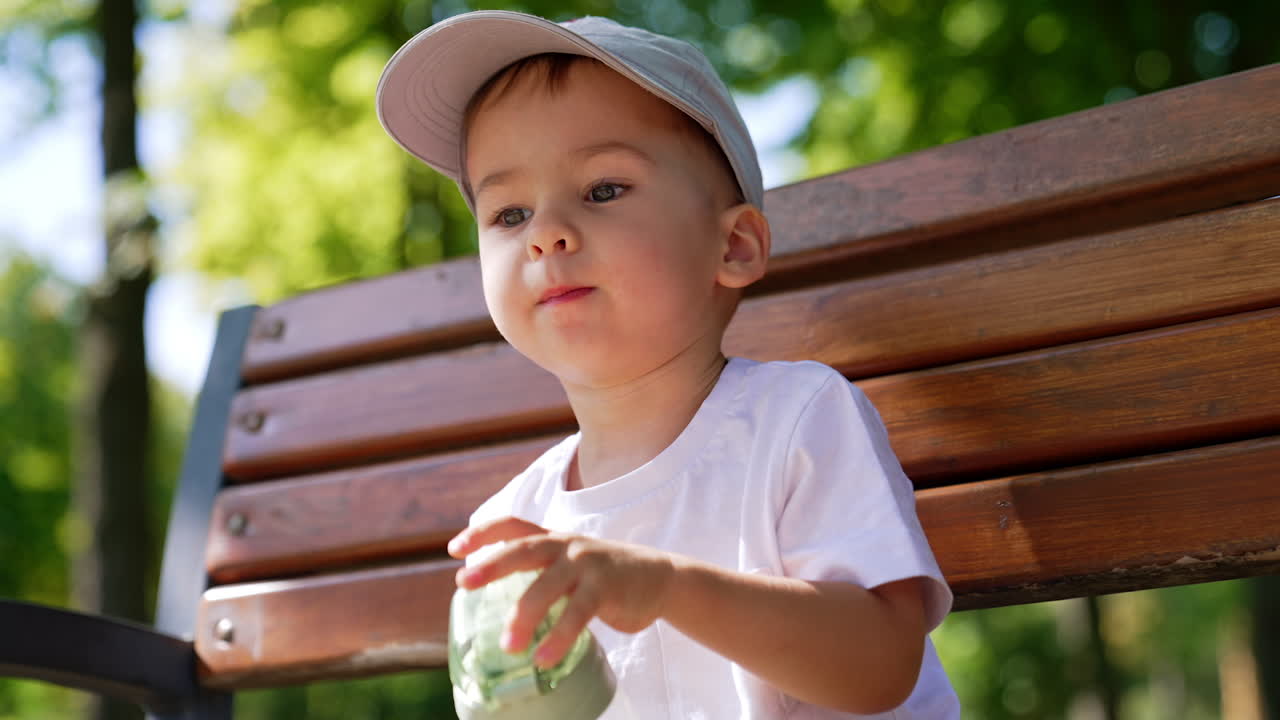 Thoughtful Caucasian baby boy in cap sitting on the bench in summer. Toddler holds the bottle with water in his hands. Low angle view.