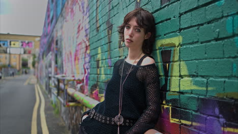 retrato de moda al aire libre de una mujer joven de estilo alternativo apoyándose en una pared cubierta de graffiti en la calle 4 de la ciudad de londres, reino unido, en tiempo real.