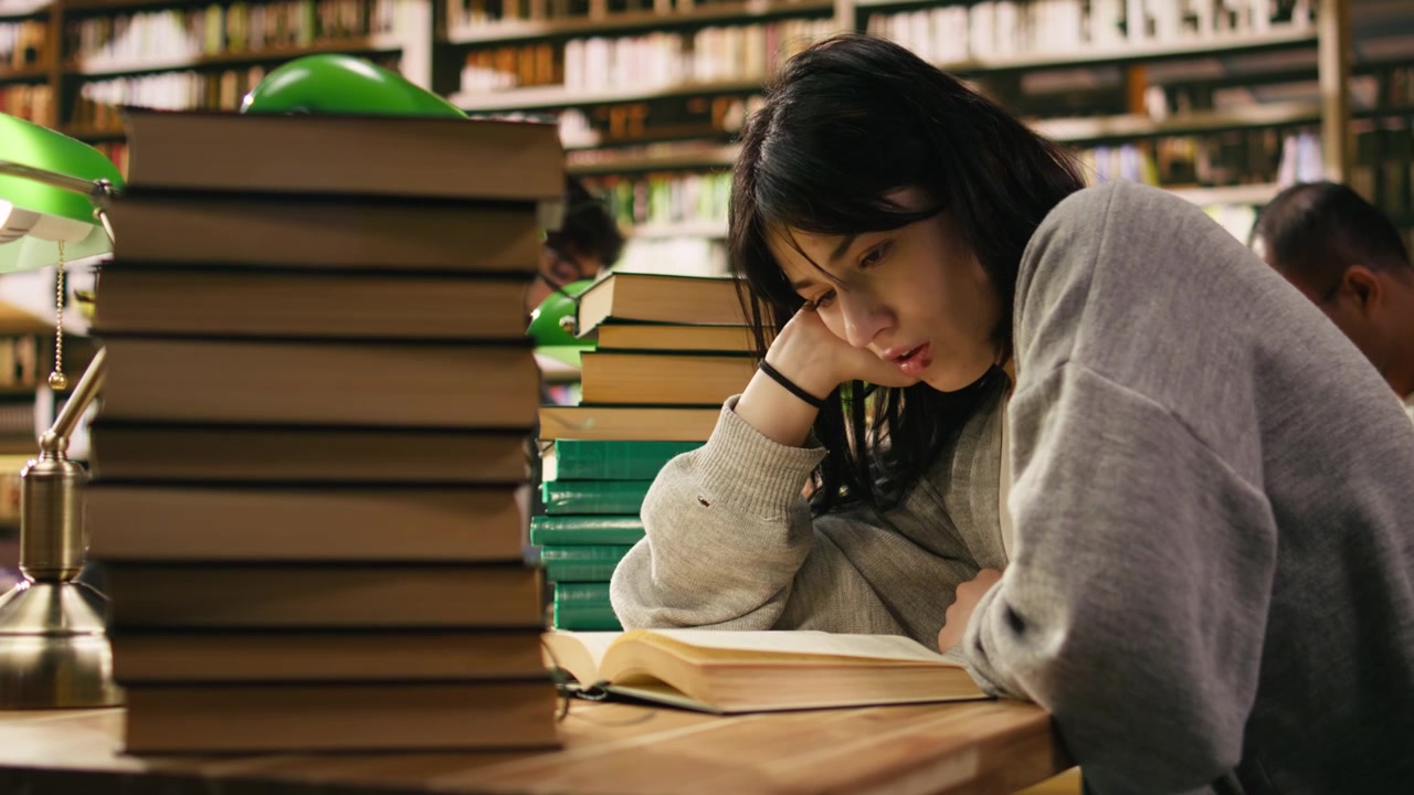 Fokusert kvinnelig student leser i åpen bok ved siden av bokstabel på biblioteket