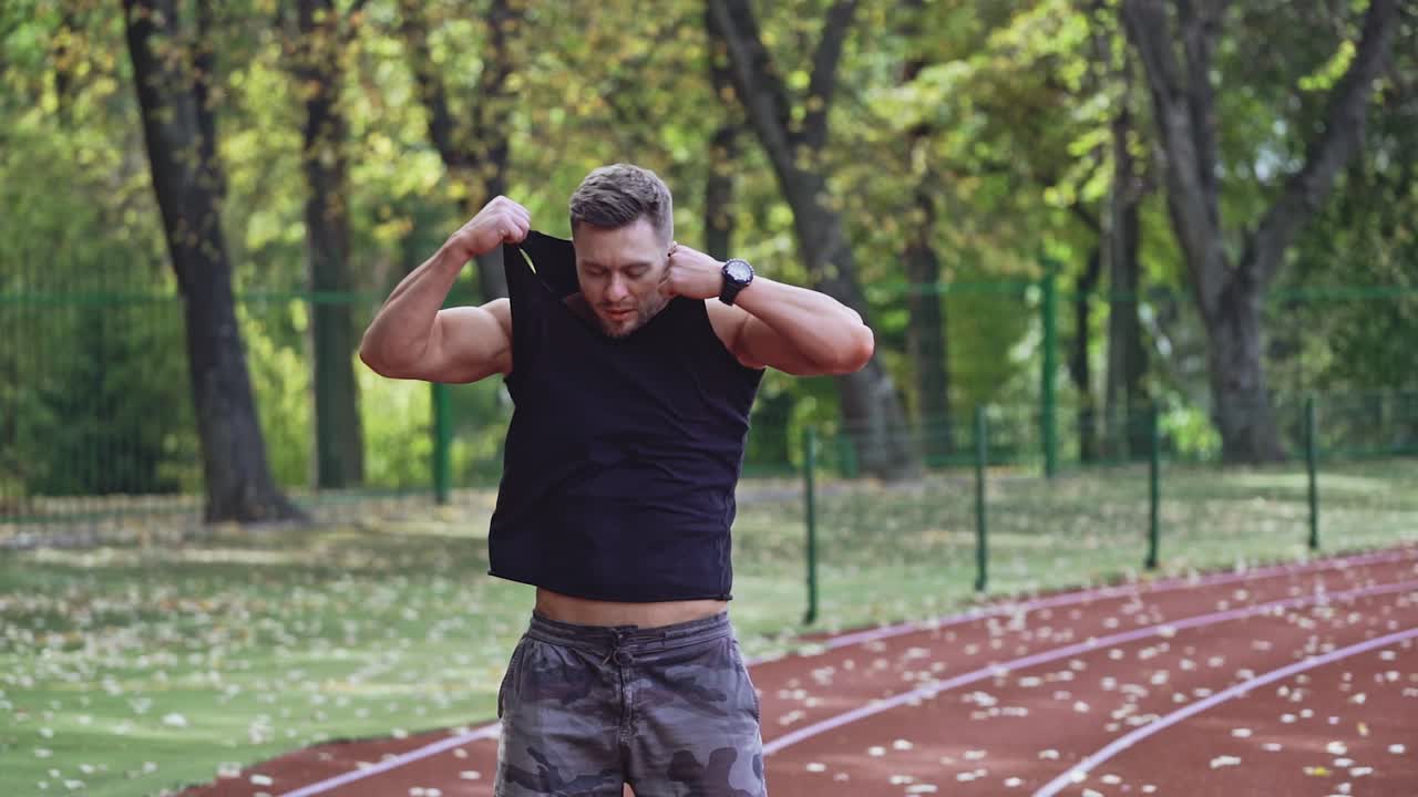 Young man takes off his jersey at the open stadium. Muscular sportsman shows his pumped body against the background of green trees.
