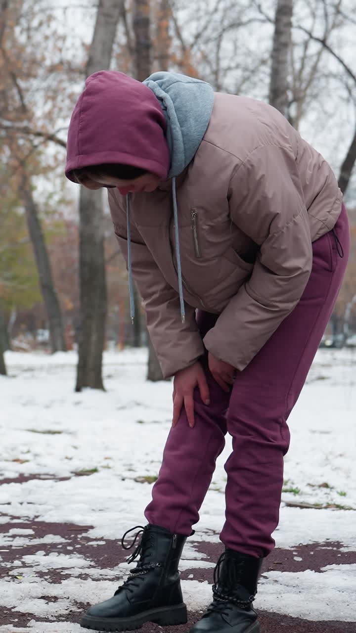 una mujer joven con traje de invierno siente dolor en la rodilla mientras se dobla y frota su rodilla derecha, con un columpio balanceándose suavemente cerca y el fondo del parque nevado