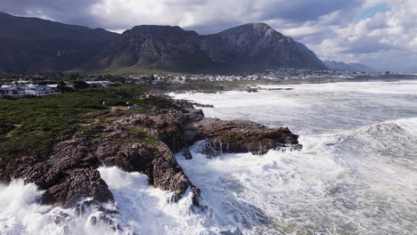 waves crash into rocky shoreline with epic mountain background, aerial tilt-up