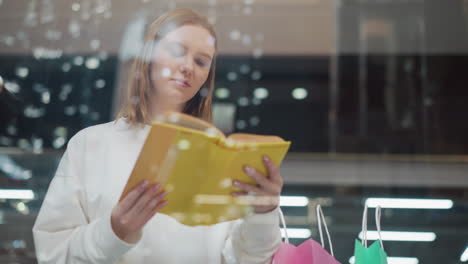 mujer joven leyendo un libro amarillo visto a través de un vidrio con reflejos de luz creando una estética moderna, bolsas de compras cercanas resaltan su comportamiento reflexivo y relajado en un entorno minorista