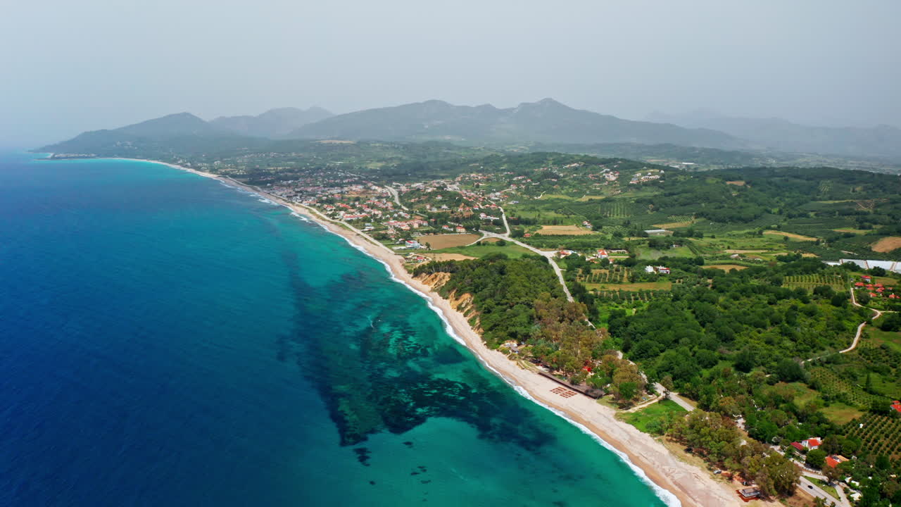 Aerial view of a picturesque coastline with a sandy beach, clear blue sea, and a town nestled against a mountainous backdrop