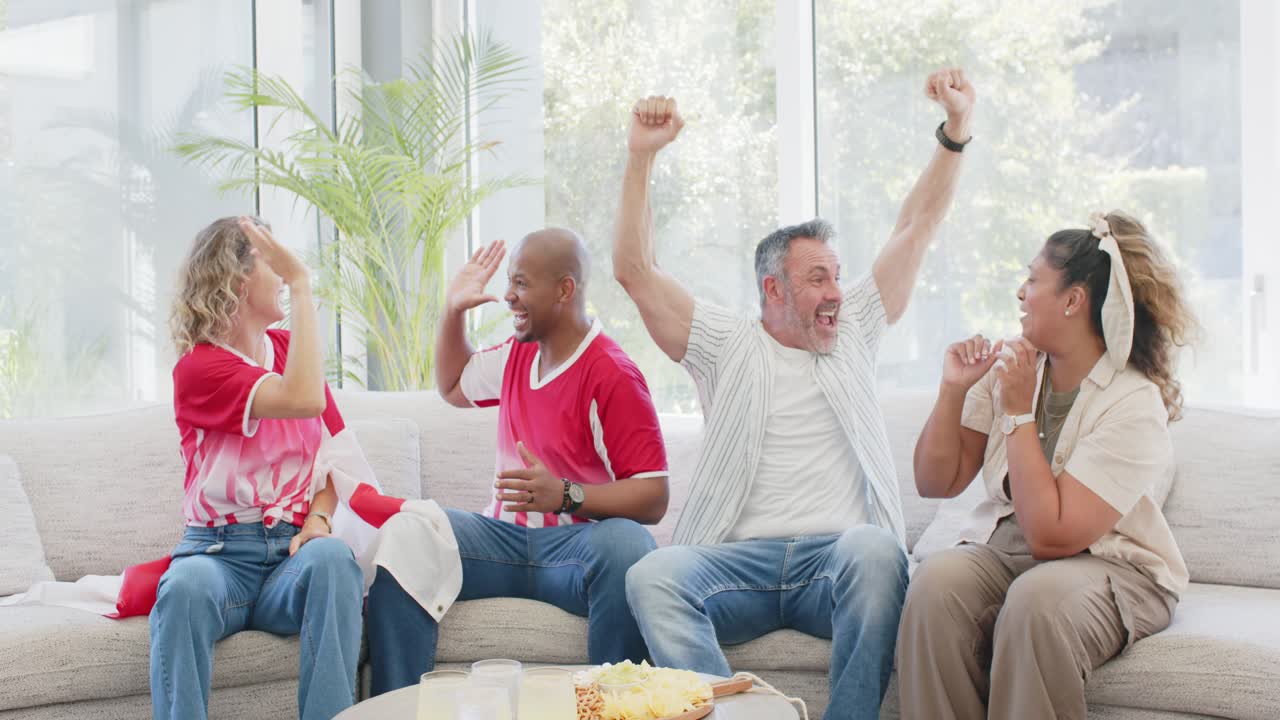 Vier Freunde in Teamshirts zu Hause auf einem geschwungenen Sofa reagieren auf ein Fernsehspiel, klatschen und geben sich High-Fives.