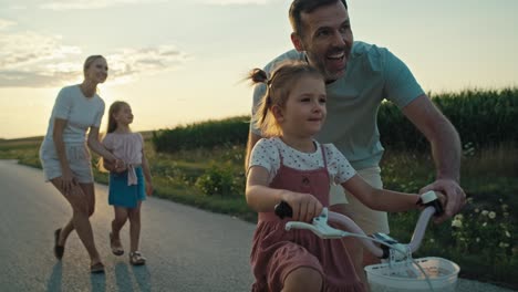 eine familie von vier weißen menschen, die bei sonnenuntergang auf der dorfstraße spazieren und fahrrad fahren.